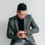 A fashionable young man in a gray suit is seated while checking his watch.